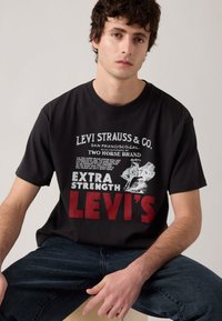 Jaunas vyras, vilkintis juodą "Levi's" marškinėlį su baltu ir raudonu grafiniu tekstu bei tamsiomis džinsomis, sėdintis prieš neutralų šviesų foną.