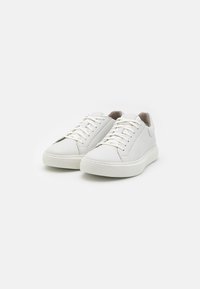 White leather sneakers with round toe, textured surface, and minimalistic design. Features flat, light-colored laces and a thick rubber sole.