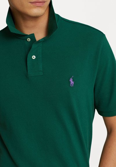 Roheline polo särk, mis on valmistatud tekstuurilisest kangast, omab kahe nupuga kaelust ning vasakul rinnal on lillakate tikandiga logo.
