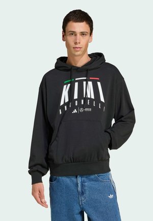 Sudadera negra con capucha y letras blancas, con acentos en verde y rojo, que presenta un bolsillo frontal y puños acanalados, combinada con unos vaqueros de mezclilla azul claro.
