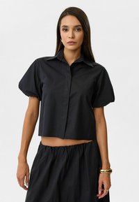 Zwarte cropped blouse met korte puffmouwen, knoopsluiting aan de voorkant en standaard kraag. Het materiaal heeft een gladde textuur en een lichte uitstraling.