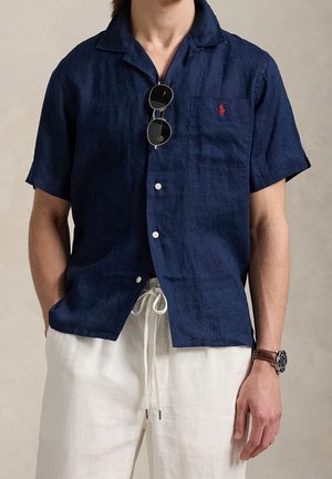 Mann trägt ein marineblaues kurzärmeliges Hemd mit rotem Logo, runde Sonnenbrille am Kragen, weiße Hose mit Kordelzug und Armbanduhr.