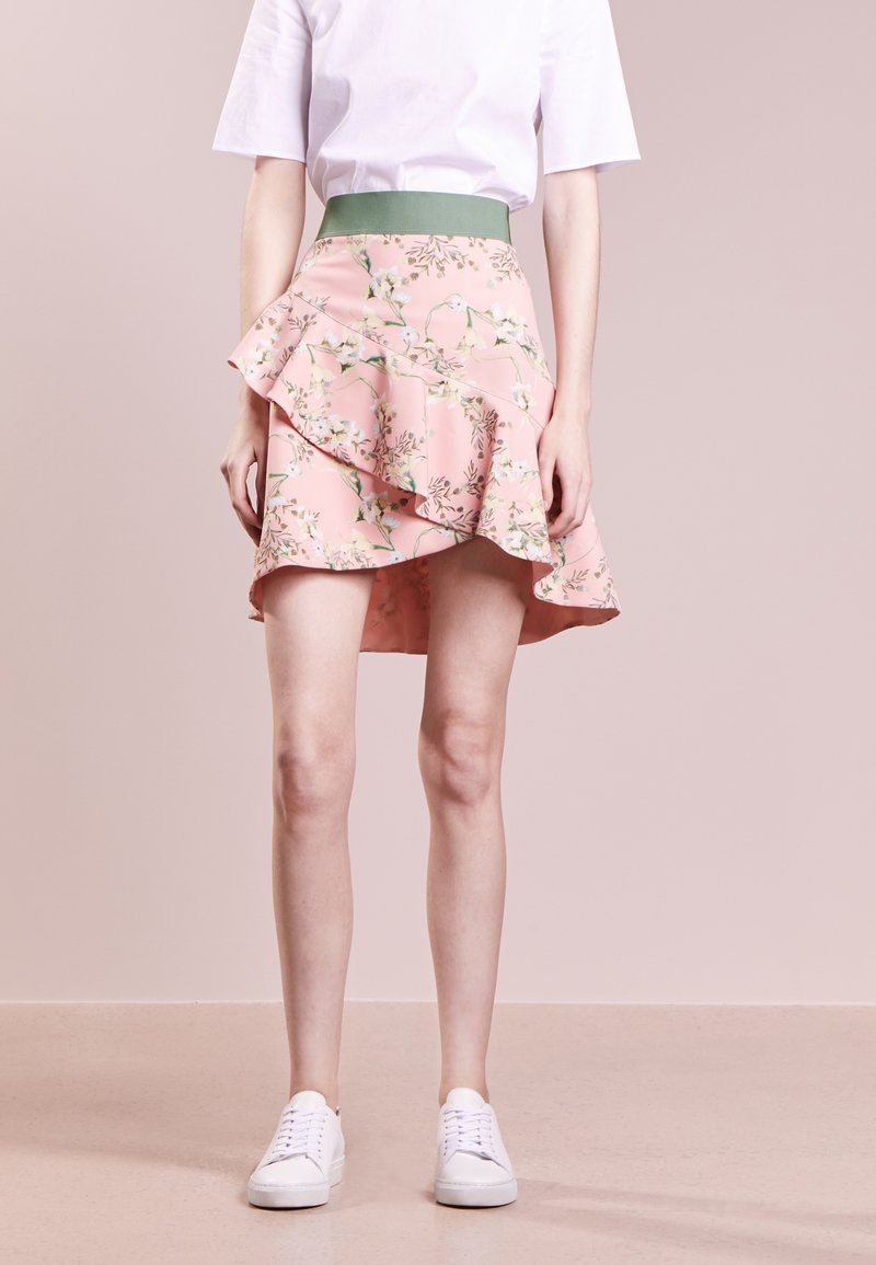 Žena v růžové květované sukni se volány a zeleným páskem, bílém tričku a bílých teniskách, stojící na béžové podlaze.