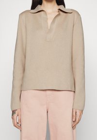 Beige lange mouwen coltrui gedragen met lichtroze hoge taillebroek, tatoeage zichtbaar op de linkerhand.