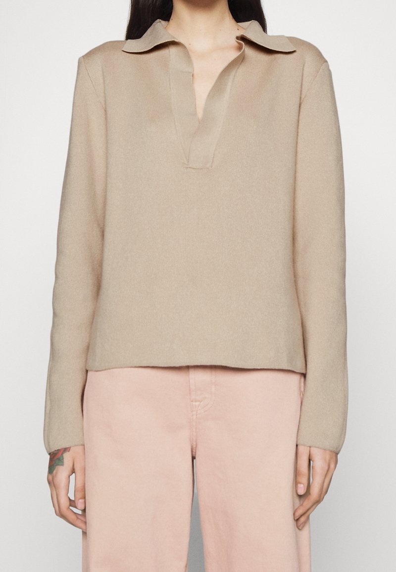 Beige lange mouwen coltrui gedragen met lichtroze hoge taillebroek, tatoeage zichtbaar op de linkerhand.