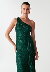 Smaragdgroene, een-schouder paillettenjurk met een aansluitende top en een gerimpeld detail aan de zijkant, die een glanzende textuur en decoratieve accenten toont.