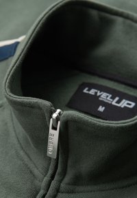 Col zippé vert foncé avec tirette argentée gravée "OMBRE" et une étiquette de taille noire indiquant "LEVEL UP M" à l'intérieur.