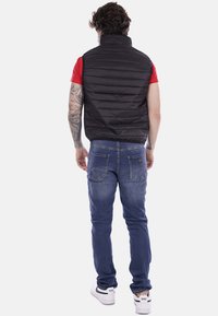 Gilet imbottito nero, colletto alto, design trapuntato. Indossato sopra una camicia rossa, abbinato a jeans blue e sneakers bianche, visto da dietro.