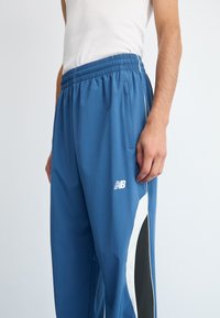 Blue athletic pants with elastic waistband, side pockets, white and black side panels, and New Balance logo on left thigh.