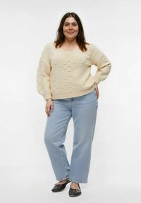 Femme se tenant debout, portant un pull en crème texturé, un jean bleu clair et des ballerines noires, devant un fond blanc uni.