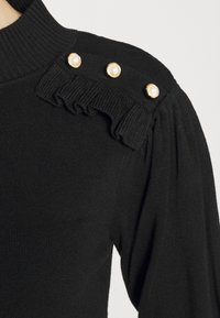 Czarny sweter prążkowany z wysokim kołnierzem, z trzema złotymi guzikami z perłami na ramieniu oraz małym detalem falbanki pod nimi.