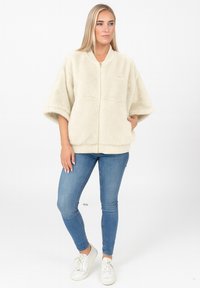 Veste en fausse fourrure couleur crème avec fermeture éclair à l'avant, manches larges et ourlet côtelé, associée à un jean skinny bleu et des baskets blanches.