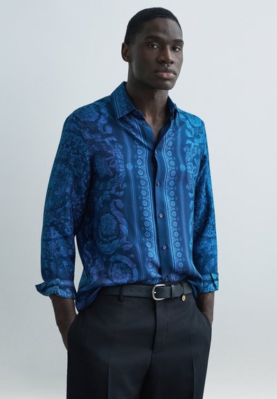 Blaues Hemd mit Muster, verziert mit filigranen Designs und einem glänzenden Finish, kombiniert mit dunkler Hose. Verfügt über Knöpfe und einen klassischen Kragen.