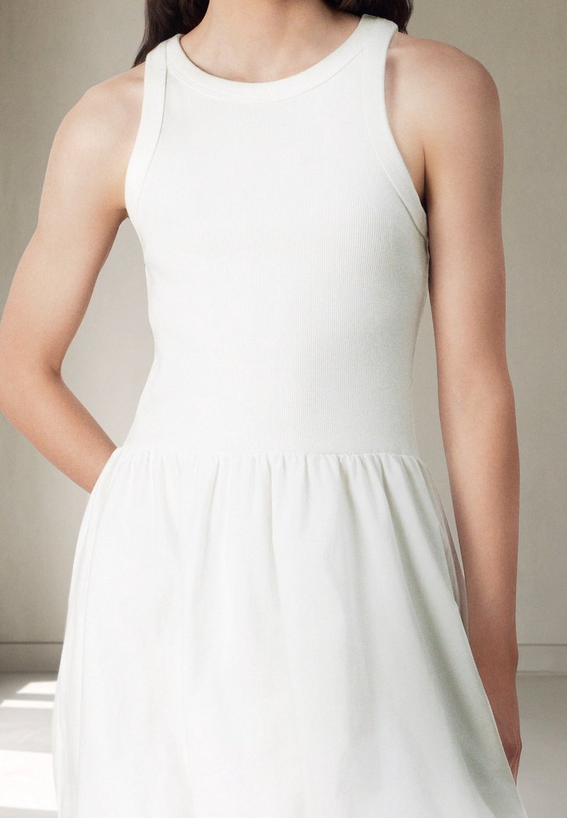 Robe blanche sans manches avec un corsage ajusté en côtes et une jupe évasée ornée de plis à la taille. Texture douce et design simple.