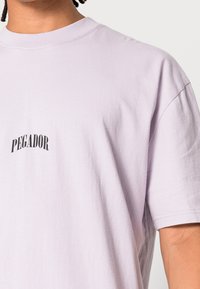 Detailní záběr na osobu, která má na sobě světle fialové tričko s nápisem "PEGADOR" vytištěným černým písmem na hrudi.