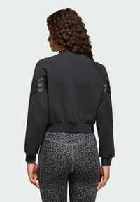 Žena s kovrčavom kosom, obučena u crnu majicu dugih rukava s prugama na rukavima i crne tajice s leopard uzorkom, gleda prema izlaznoj strani.