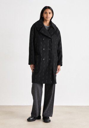 Zwarte wollen jas met grote revers, dubbelzijdig ontwerp, voorzakken en witte streepjes. Gecombineerd met grijze wijde broek met stippen.