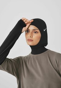 Hijab sportiv negru cu un design ajustat și o textură netedă. Asortat cu un top lejer, cu mânecă scurtă, de culoare măslinie. Logo-ul Nike este vizibil.