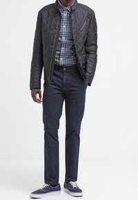 Man som bär en svart vadderad jacka över en blå rutig skjorta, marinblå byxor och marinblå sneakers med vita sulor, stående på vit bakgrund.