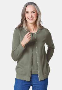 Olijfgroene cardigan met ronde hals, knoopsluiting en lange mouwen. Zachte textuur en getailleerd ontwerp, gecombineerd met blauwe spijkerbroeken.