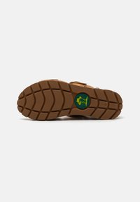 Brun teksturert såle med dype spor for grep, med en rund grønn logo med et fiskedesign i midten.