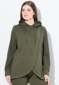 Mujer con una sudadera con capucha de color verde oliva y panel frontal cruzado, y pantalones a juego, posando frente a un fondo blanco liso.