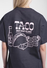 Mörkgrå bomull t-shirt med rosa grafisk design av en taco, en flaska het sås, tortillachips och glas med en citronskiva.