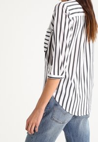 Blouse à rayures noires et blanches avec manches trois-quarts, ourlet arrondi et poche plaquée. Tissu doux à texture lisse.