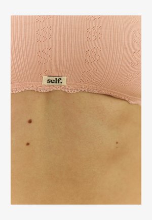 Rozā ribota krūšturis ar mežģīņu apdari, ar smalku rakstu un etiķeti "self." Novietots uz ādas fona.