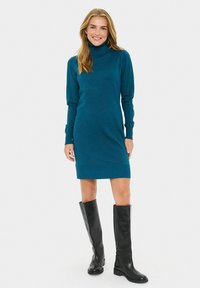 Abito a maglia turchese con collo alto, maniche lunghe, design aderente e polsini a coste, abbinato a stivali neri di pelle al ginocchio.