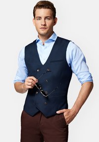 Granatowa kamizelka dwurzędowa z teksturowanej tkaniny, zestawiona z jasnoniebieską koszulą. Model trzyma okulary w jednej ręce, nosząc brązowe spodnie.