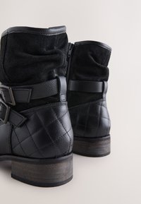 Black ankle boots featuring a combination of textured leather and suede, quilted detailing, adjustable straps, and a small wooden heel.