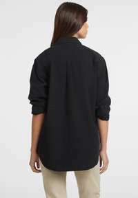 Camicia nera con bottoni, dal taglio rilassato, con maniche arrotolate e orlo arrotondato. Realizzata in un tessuto leggero con una texture semplice.