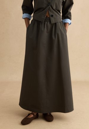 Mujer con falda larga de color verde oliva oscuro con cintura elástica, combinada con chaqueta corta a juego y zapatos planos marrones tejidos. Manos en los bolsillos.