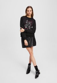 Čierna mikina s bielymi a červenými grafickými vzormi, skombinovaná s čiernou sukňou z imitácie kože. Model má na sebe čierne členkové topánky.