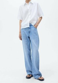 Camisa blanca de botones de manga corta con un corte relajado, combinada con jeans de pierna ancha de color azul claro. Sandalias de suela plana completan el conjunto.