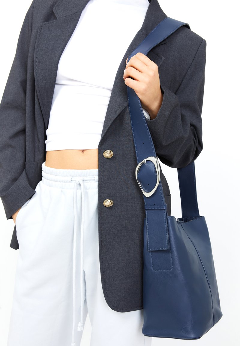 Borsa a secchiello in pelle blu navy con una larghezza della tracolla e dettagli in metallo argentato. Texture liscia, silhouette strutturata.