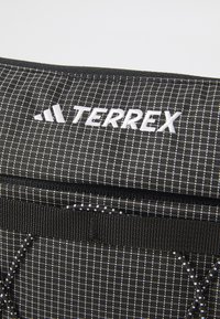 Geantă din material textil negru cu logo-ul alb TERREX. Are un model în grilă, un design plat și un sistem de închidere cu șnur pentru o prindere sigură.