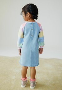 Lichtblauwe spijkerjurk met pastelkleurige raglanmouwen, voorzien van knoopdetails aan de achterkant. Gecombineerd met glinsterende sneakers en roze gerimpelde accenten.