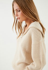 Kvinna med långt blont hår som bär en krämfärgad, texturerad stickad tröja med bred krage, som tittar nedåt och åt sidan mot en enfärgad bakgrund.