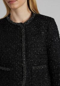 Schwarze strukturierte Jacke mit Rundhalsausschnitt, verziert mit Perlen, ausgestattet mit zwei Fronttaschen und dekorativer Hardware.