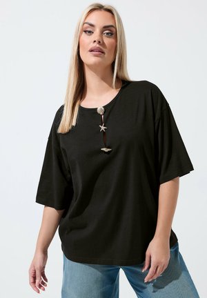 Blonde Frau trägt locker sitzendes schwarzes T-Shirt mit drei muschelförmigen Knöpfen und hellblaue Jeans, steht vor einfachem weißem Hintergrund.