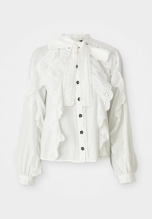 Hvit bluse med lange, puffede ermer og krage med sløyfe. Har skjellkantede kanter og broderte blomsterdesign langs fronten. Svart knapper.