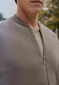 Light brown zip-up sweater with a textured fabric, ribbed collar, and subtle stitching details, layered over a beige shirt.