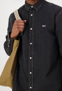 Mörk denimskjorta med knappknäppning, kontrasterande sömmar och en liten logotypmärke; hålls med en beige tote-väska.