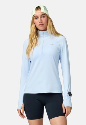 Mladá žena v svetlomodrom športovom tričku s dlhým rukávom, čiernych šortkách, svetlozelenom šiltovke nasadenej naopak a so smart hodinkami na ľavom zápästí.