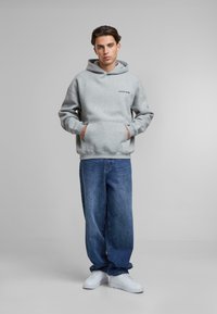 Hanorac gri cu glugă și buzunar frontal, având un logo minimalist de culoare neagră. Purtați cu blugi largi albaștri și adidași albi.