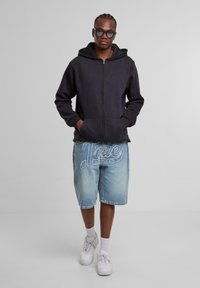 Joven vestido con una sudadera con cremallera negra, grandes gafas de sol tintadas, pantalones cortos de mezclilla con bordado blanco, calcetines blancos y zapatillas blancas, de pie contra un fondo gris.