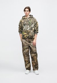 Camo hoodie and pants set in earthy tones with leaf and bark patterns; features a front pocket and ribbed cuffs. White sneakers complete the look.