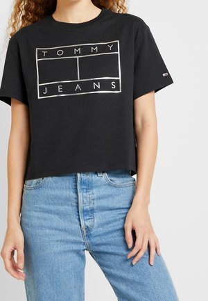 Person iført en sort cropped T-shirt med "Tommy Jeans" logo og lyseblå højttaljede jeans, stående mod en ensfarvet baggrund.
